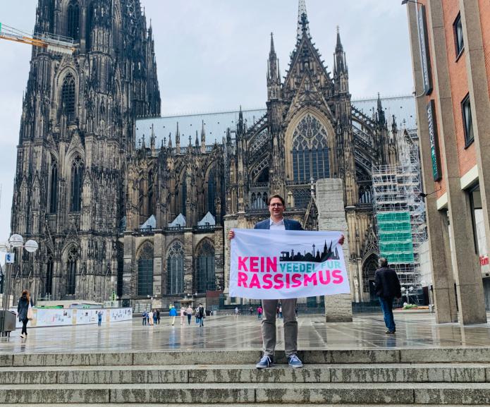 Maximilian Rudloff vom Vorstand des Katholikenausschusses in der Stadt Köln mit einer Aktionsfahne gegen Rassismus. Foto: © Katholikenausschuss in der Stadt Köln  